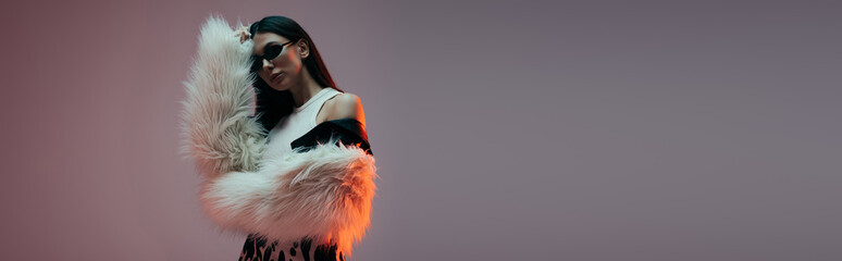 stylish young woman in sunglasses and white faux fur jacket posing on grey, banner.