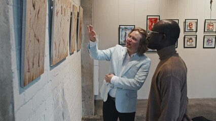 Medium long side view of blond-haired Caucasian man and his young African American friend discussing series of paintings in modern art gallery at daytime