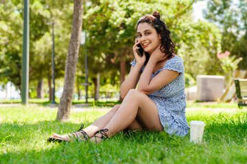 Cheerful brunette woman wearing summer dress on city park, outdoors talking on mobile phone with friends or boyfriend with smiles. Looking at the camera.