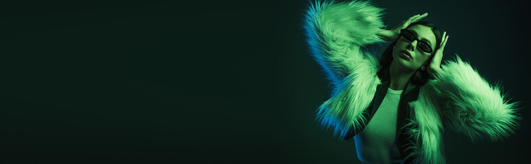 stylish young woman in faux fur jacket and sunglasses posing on dark background, banner.
