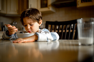 A five-year-old boy with a dissatisfied face has breakfast at home in the kitchen, the child eats porridge and drinks milk