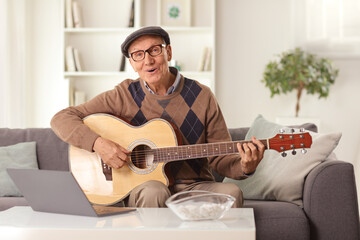 Elderly man playing an acoustic guitar and sitting on a sofa in front of a laptop computer