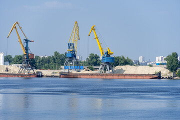 Fototapeta na wymiar Big port cranes on the river. Extraction and transportation sand. Harbor crane loads water resources into the barge. Special equipment and water transport. River port. Industrial city.