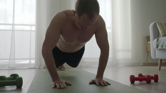 4K Man doing push-up exercises at home as part of his work-out session. Man working out on yoga mat with dumbbells sitting at both sides, sports and healthy lifestyle concept.