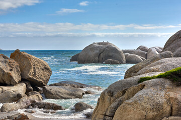Beautiful sea view of big boulders and ocean water on a sunny beach day in summer. A seaside view of nature with a blue sky, white clouds, and waves. A seascape near the shore under the horizon