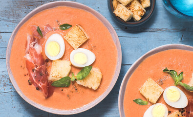 Spanish salmorejo soup, tradition Andalusian cold tomato soup topped with serrano ham (jamon), boiled egg, bread and pepper on the blue background. Summer cold recipes

