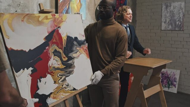 Medium long of blond Caucasian man in formal suit leading auction, workmen bringing out abstract painting