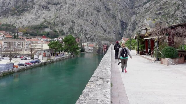 Young curvy woman tourist with backpack walks on sidewalk past river against giant mountains IN old town Kotor, Montenegro