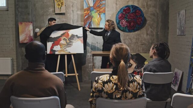 Rear view of diverse buyers sitting on chairs, looking at young Caucasian man in formal suit leading auction, workman taking black cloth off of contemporary abstract painting