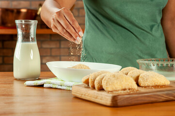 Woman breading brazilian meat stuffed croquette (risolis de carne) on a wooden table. Top view.