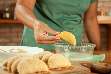 Woman preparing brazilian meat stuffed croquette (risolis de carne) with milk and breadcrumbs on a wooden table.