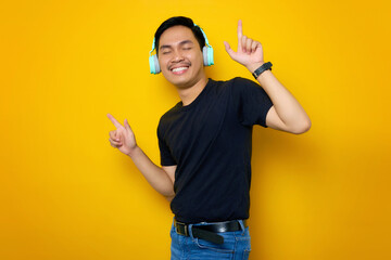 Cheerful young Asian man in casual t-shirt listening music in headphones and dancing isolated on...