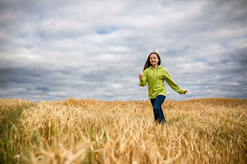 Naklejka na ściany i meble Beautiful smiling preteen girl running against cloudy sky and wheat field in summer