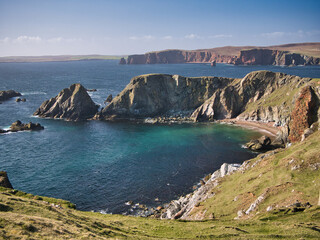 Dramatic coastal cliff scenery and a remote inaccessible beach on the Ness of Hillswick, Northmavine, in the UNESCO Global Geopark of Shetland, UK - taken on a sunny day showing the clear, blue water. - obrazy, fototapety, plakaty