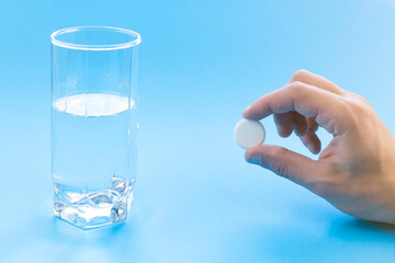 A male hand holds a pill near a glass of water on a blue background.