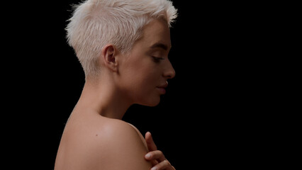 Half-turned good-looking young slim Caucasian woman with short light hair touches her arm on black background | Body care concept