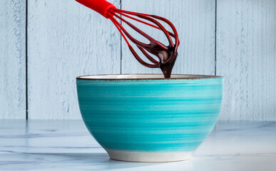 Teal blue bowl on a light table and background with a red whisk dripping melted chocolate 