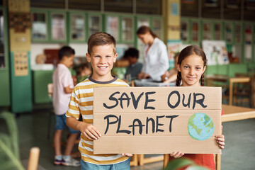 Happy elementary students holding placard with 'save our planet' message in  classroom and looking...