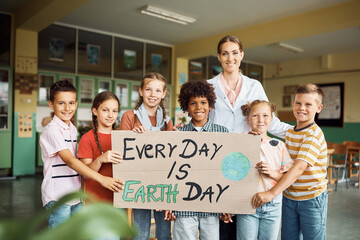 Science teacher her elementary students holding placard with 'every day is Earth day' message at school and looking at camera.