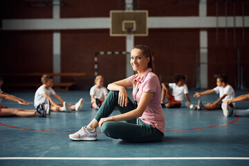 Happy physical education teacher with group of students in background at school gym.