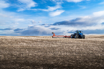 An eight wheel tractor pulling a plow across a tilled field with rolling hills in Rocky View County Alberta Canada under a dramatic cloudy sky.