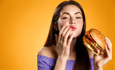 Chewing woman holding tasty fast food burger, cleaning her mouth after taking a bite. Satisfied girl eats her delivery meal, orange background