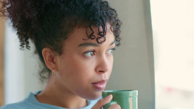 Face of beautiful african woman blowing her hot coffee or tea while looking out the window and relaxing at home. Serious woman thinking and daydreaming about her life and future after the pandemic