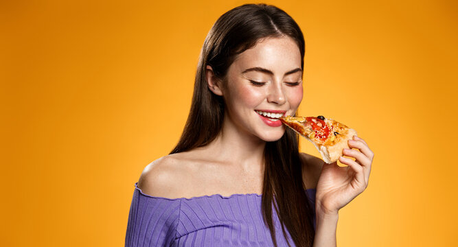 Image of girl smiling while biting slice of pizza, happy face at pizzeria restaurant, orange background
