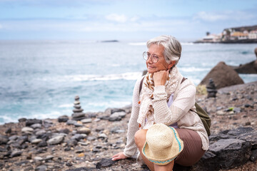 Attractive relaxed senior woman sitting on a pebble beach looking away pensive, elderly caucasian female enjoying travel vacation retirement