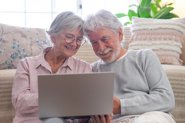 Smiling beautiful caucasian senior couple sitting on the floor at home using laptop, modern retired elderly people surfing the net with computer