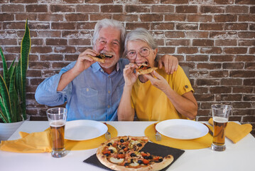 Cheerful senior couple having fun together eating a slice of pizza. Sitting at home table with beer...