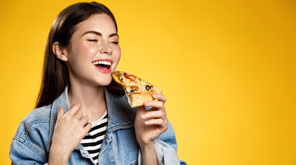 Image of girl smiling while biting slice of pizza, happy face at pizzeria restaurant, yellow...