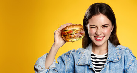 Happy smiling woman posing with cheeseburger, holding big tasty burger, enjoyment face, yellow background