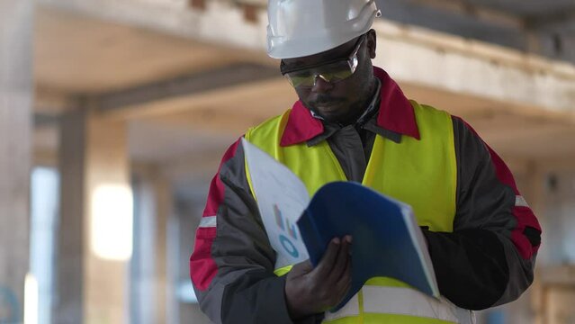 African american worker in hard hat and protective goggles stands at construction site, looks in work papers