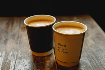 Two disposable cups of coffee with the heart on foam on a wooden table.