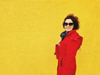 Caucasian woman wearing a red coat and sunglasses smiling in front of a yellow wall - Powered by Adobe