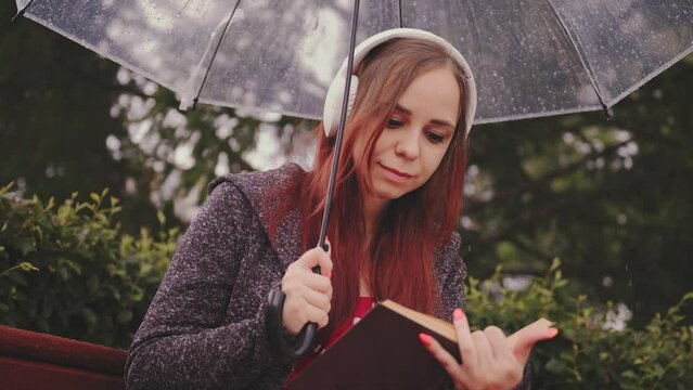 Young Beautiful Woman In Headphones With Transparent Umbrella Reading Book, Sitting On Bench In City Park In Rain. Relaxed Secluded Female Relaxing With Book, Music Sheltering With Umbrella From Rain