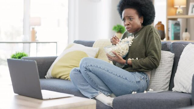 Woman watching spooky horror movie on a laptop and eating popcorn while sitting on the couch at home. Terrified african afro female enjoying her online subscription and streaming scary films online