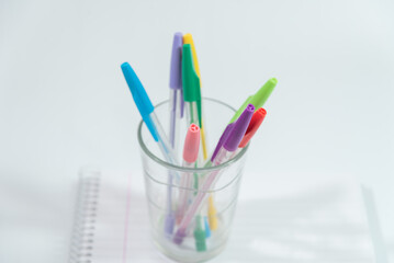pens of different colors in a glass glass over a blank notebook