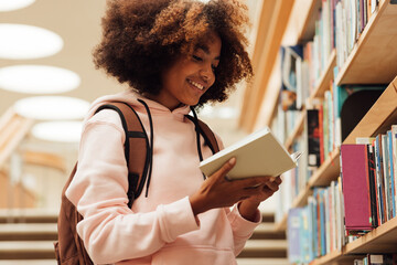 Smiling girl holding a book in library while standing at bookshelf - obrazy, fototapety, plakaty