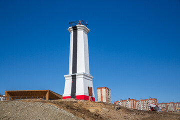 Lighthouse (leading beacon) on the shore of the sea bay in the port town. White lighthouse and residential buildings against the blue sky. Mayak Park in Magadan. The Russian Far East.