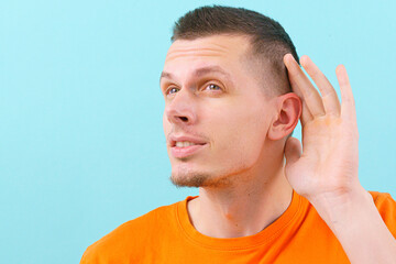 A young man in an orange shirt holding his hand near his ear and listening carefully over the blue...