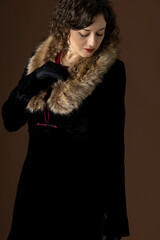 A 1920s woman wearing a black velvet coat with a fur collar and standing against a plain studio backdrop