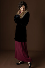 A 1920s woman wearing a black velvet coat with a fur collar and standing against a plain studio...