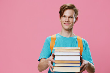a red-haired student guy in a blue T-shirt and with an orange backpack on his back, holds a heavy stack of books in his hands, turning his face away from the camera. Horizontal photo with empty space