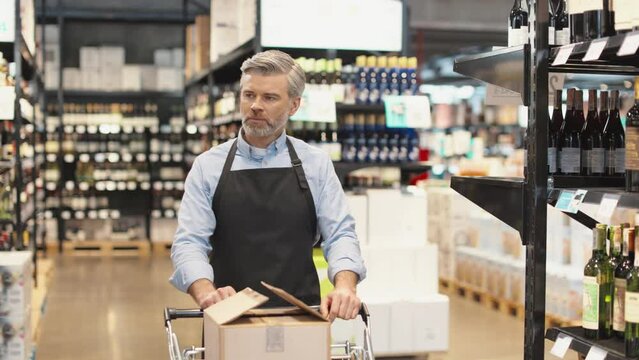 Male shopkeeper in apron pulling shopping cart with boxes of wine while walking between rows of shelves in liquor store department. Concept of people and occupation.