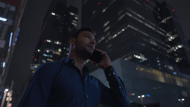 Businessman picks up a smartphone call and talk on phone near office at night, successful employee standing near skyscraper building talking on cellphone
