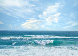 Landscape banner view of waves rolling onto the seashore against a cloudy blue sky in summer....