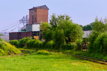 Fototapeta na wymiar Morning landscape on the railway tracks. Sunbeams and a clearing of green grass between the rails. Red brick tower and trees along the railway in the suburbs of the industrial area. Summer greens.