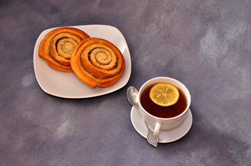 Two fresh cinnamon rolls and a cup of hot tea with a slice of lemon on a gray background.
